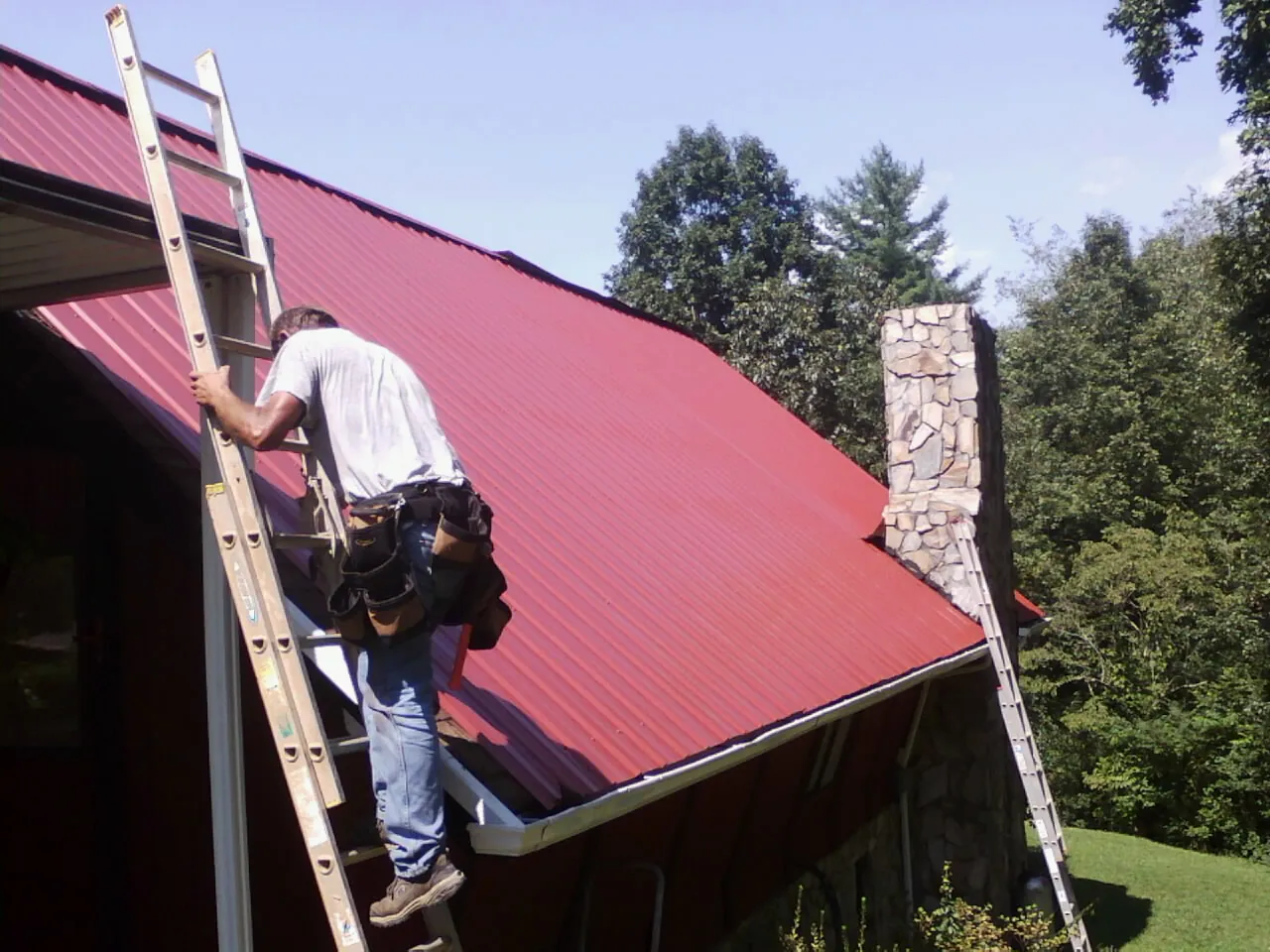 Roofer on a ladder beside a red metal roof and stone chimney — Soffit & Fascia Repair by Roofing Brothers in Castle Dale